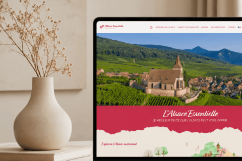 Un ordinateur portable posé sur une table beige affiche le site web d'Essentielle, sur lequel figure une photo panoramique d'un village et d'une église en Alsace, en France. Un vase aux tons neutres, contenant des branches séchées, est posé à côté de l'ordinateur portable, devant un mur texturé.