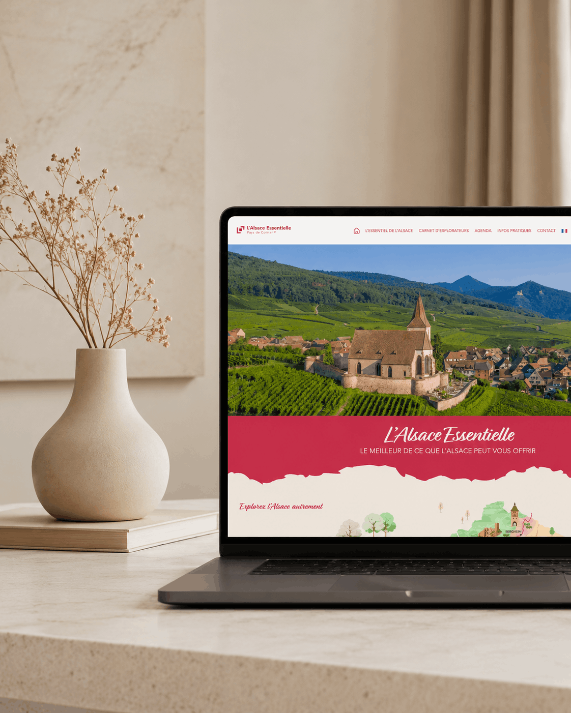 Un ordinateur portable posé sur une table beige affiche le site web d'Essentielle, sur lequel figure une photo panoramique d'un village et d'une église en Alsace, en France. Un vase aux tons neutres, contenant des branches séchées, est posé à côté de l'ordinateur portable, devant un mur texturé.
