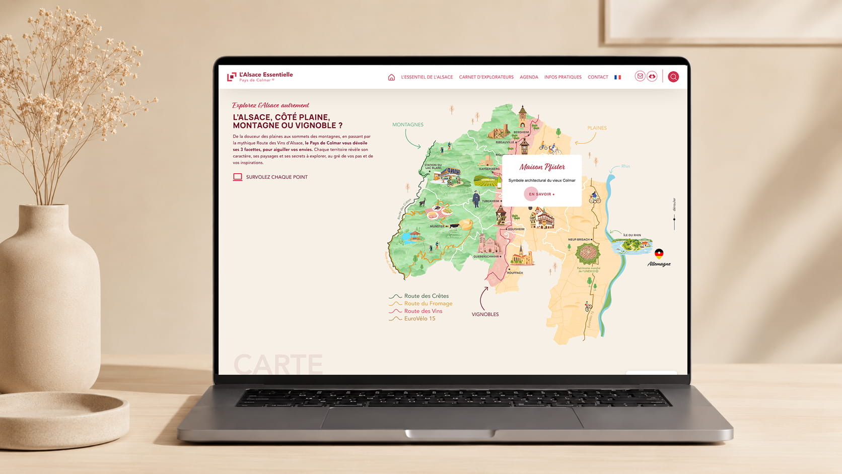 Un ordinateur portable posé sur un bureau beige affiche une carte illustrée et colorée de l'Alsace, en France, mettant en évidence les principales attractions touristiques. La carte est présentée sur un site web dont la partie supérieure comporte des menus de navigation. Des objets de décoration sont disposés à côté de l'ordinateur portable.
