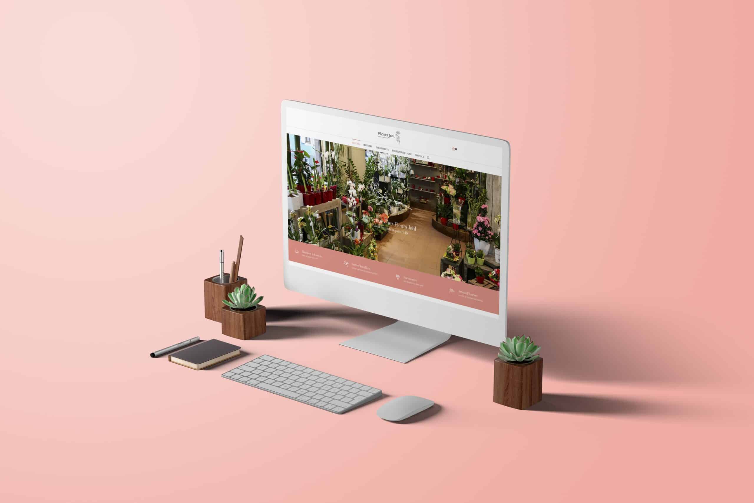 Un ordinateur de bureau affichant le site web d'un magasin de fleurs est posé sur une surface rose, entouré d'un clavier, d'une souris, d'un bloc-notes, d'un porte-stylo et de deux petites plantes grasses en pot.