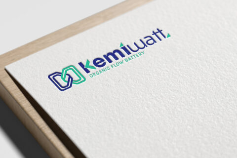 Gros plan sur un papier blanc texturé, sur lequel sont imprimés le logo bleu et vert de Kemiwatt ainsi que la mention « Kemiwatt Organic Flow Battery », posé sur une surface en bois.