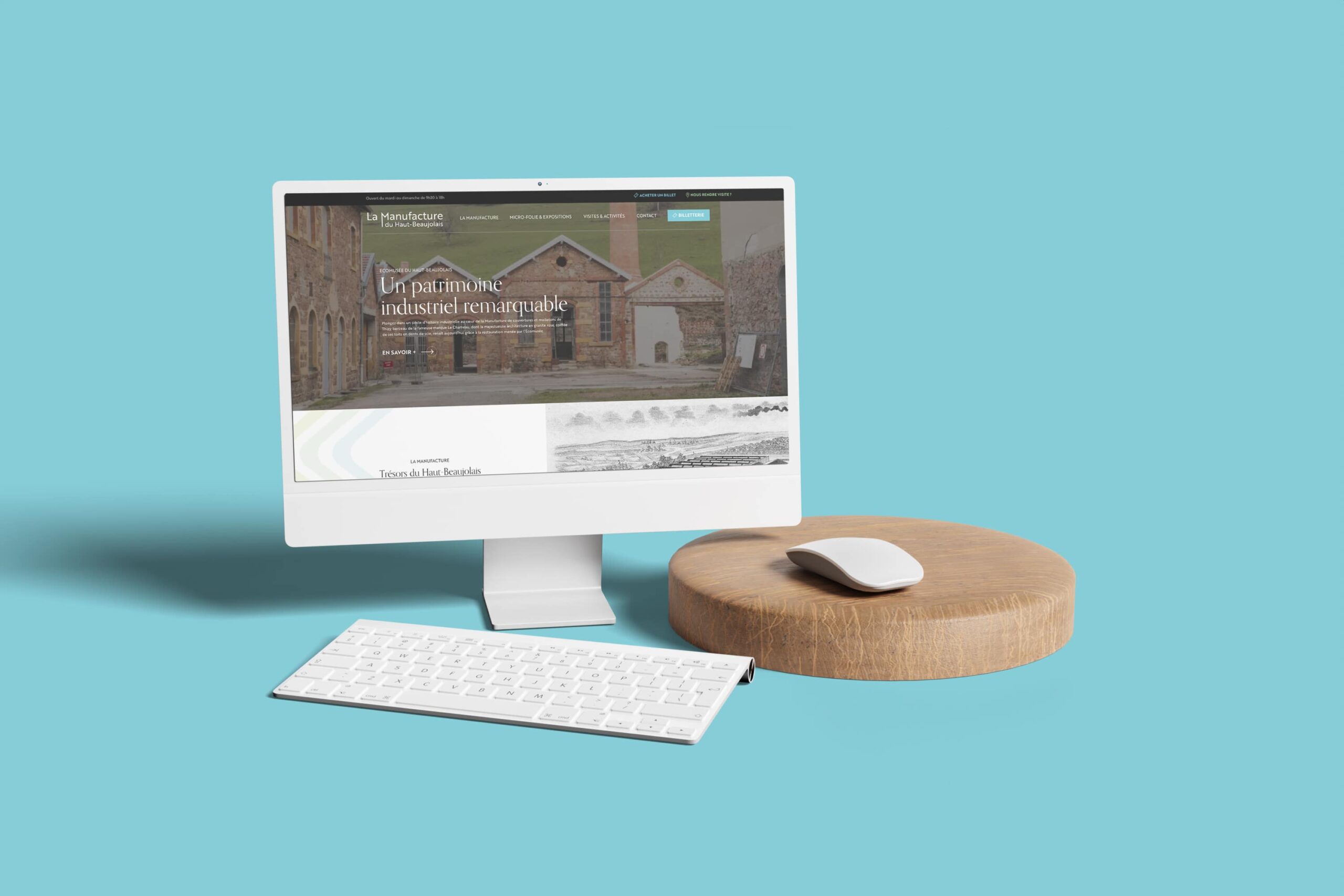 Un ordinateur de bureau blanc avec un clavier et une souris blancs se trouve sur un fond bleu clair. L'écran affiche un site web pour La Manufacture, avec des bâtiments historiques et le texte "Un patrimoine industriel remarquable".
