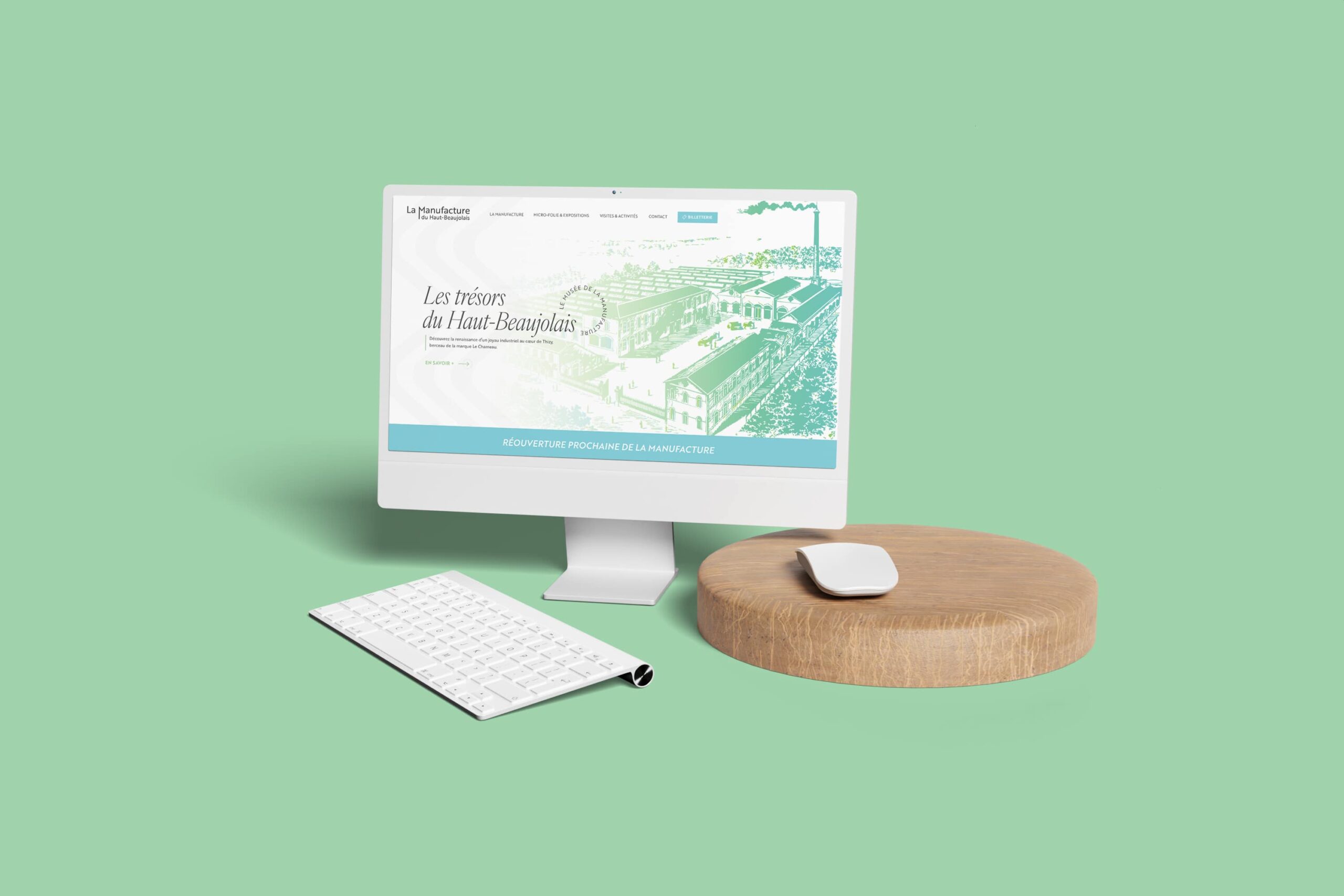 Un ordinateur blanc affichant le site Internet de La Manufacture, accompagné d'un clavier et d'une souris assortis. La souris repose sur un support rond en bois, le tout disposé sur un fond vert pastel.